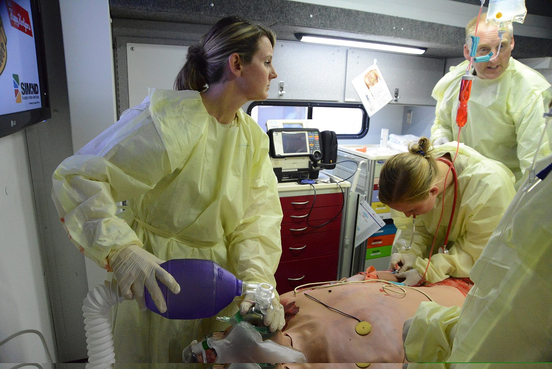 Instruktörer och deltagare tränar vid en patientsimulator i ett vårdrum.